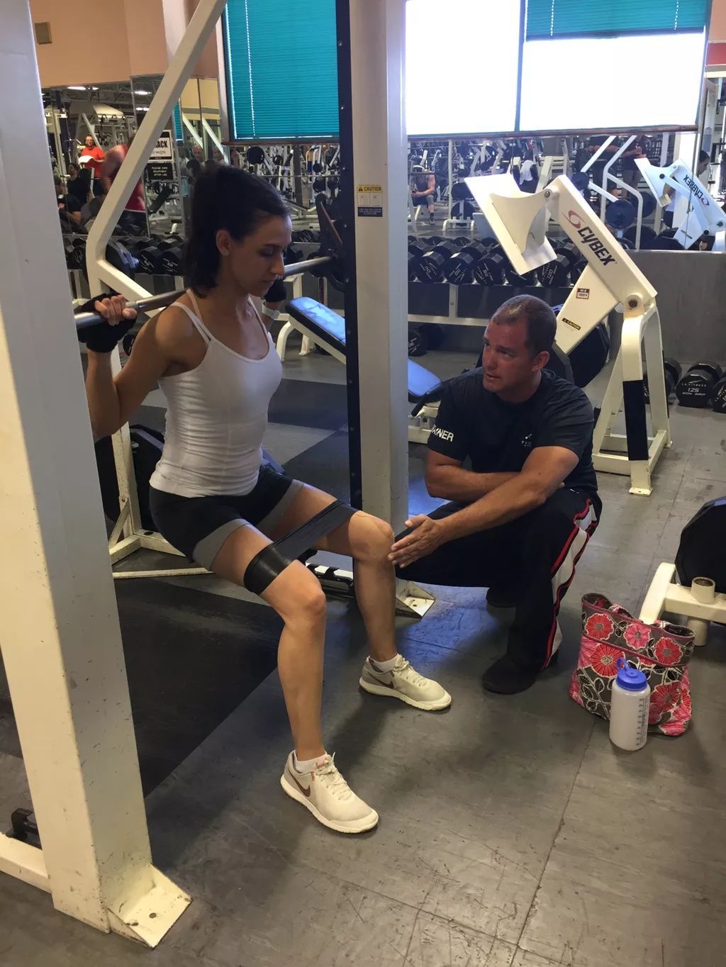 Person doing a wall sit exercise in a gym while a trainer kneels nearby.