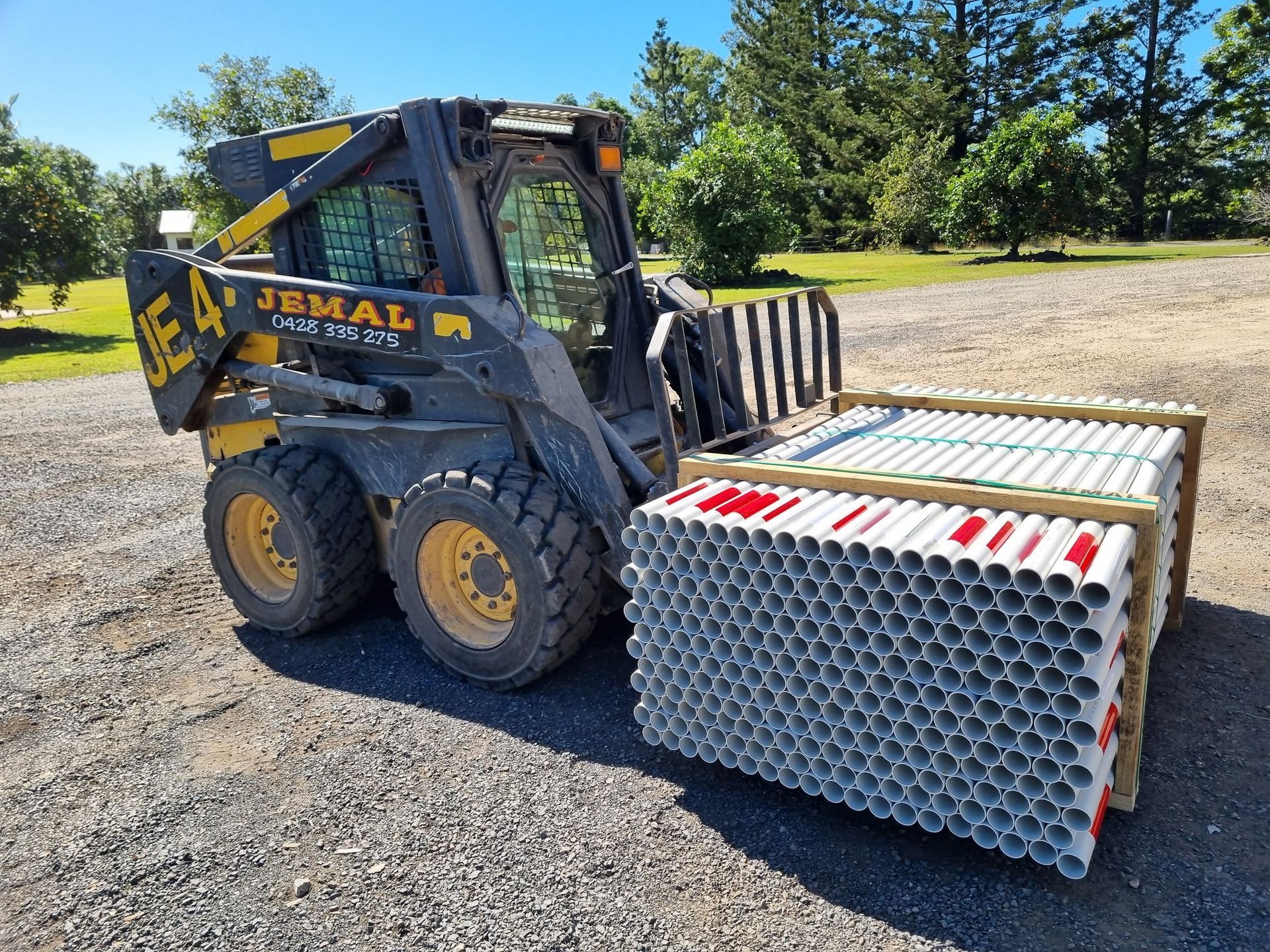 Skid steer loader carrying a pallet of numerous white and red plastic tubes outdoors on gravel — Jemal Enterprises In Finch Hatton, QLD