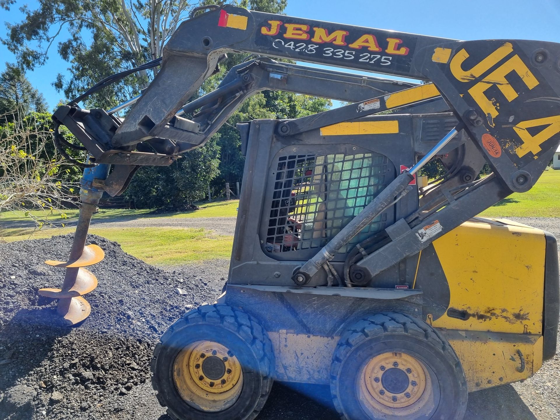 Skid steer with auger drilling into a pile of dark gravel, outdoors.