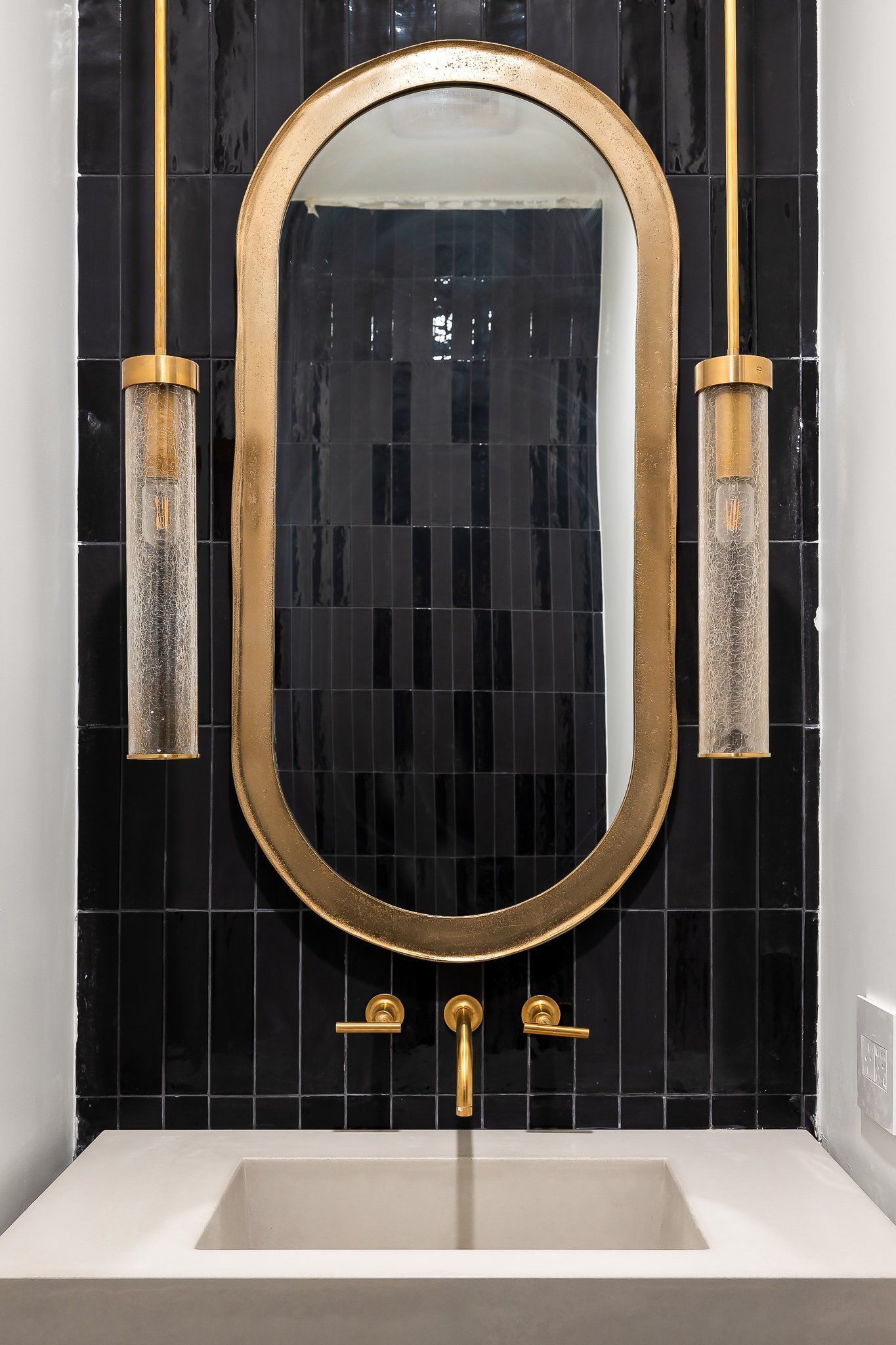Gold-framed oval mirror above a sink, flanked by two pendant lights against glossy black tile.