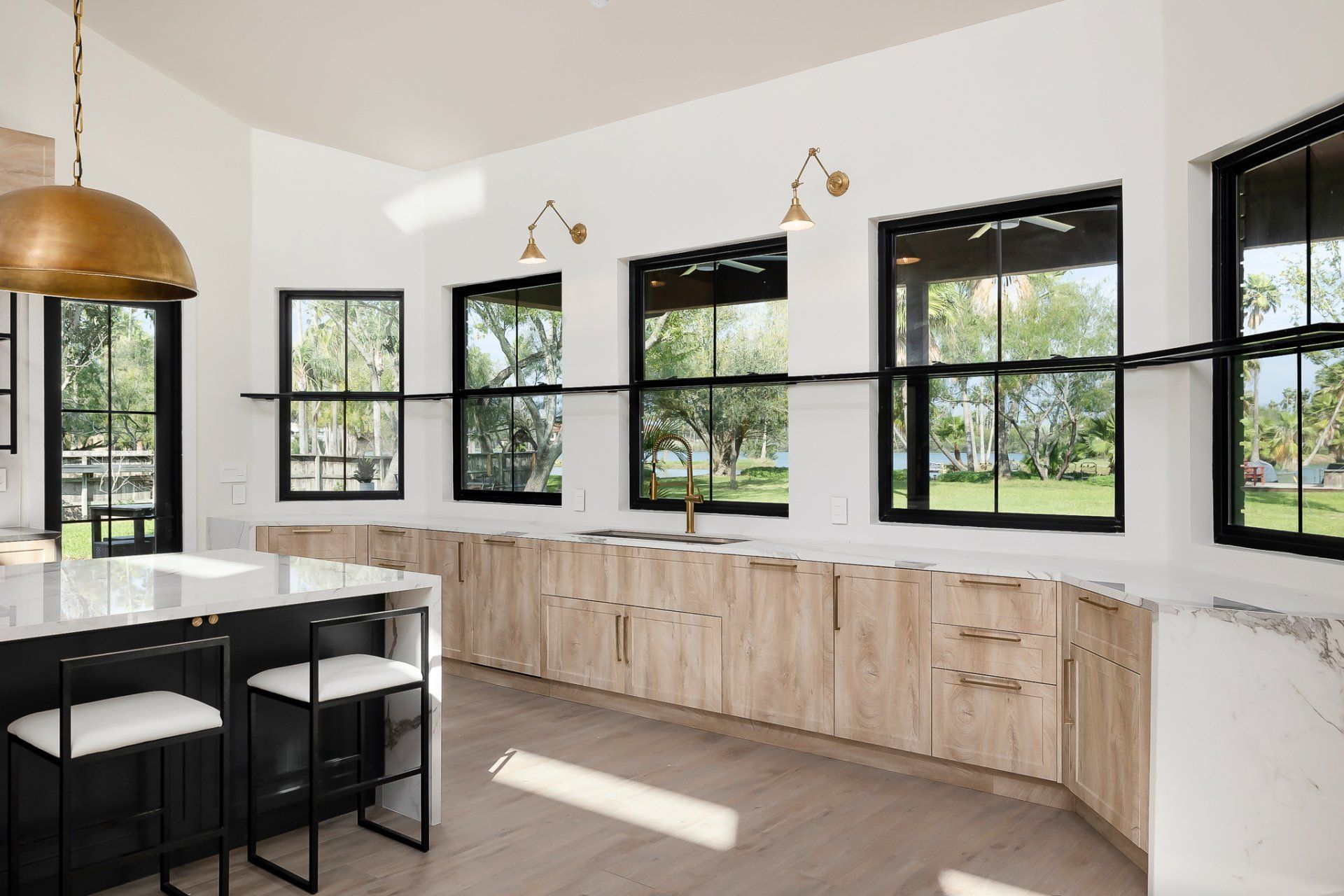 Modern kitchen with black-framed windows, light wood cabinets, marble countertops, and brass pendant lights