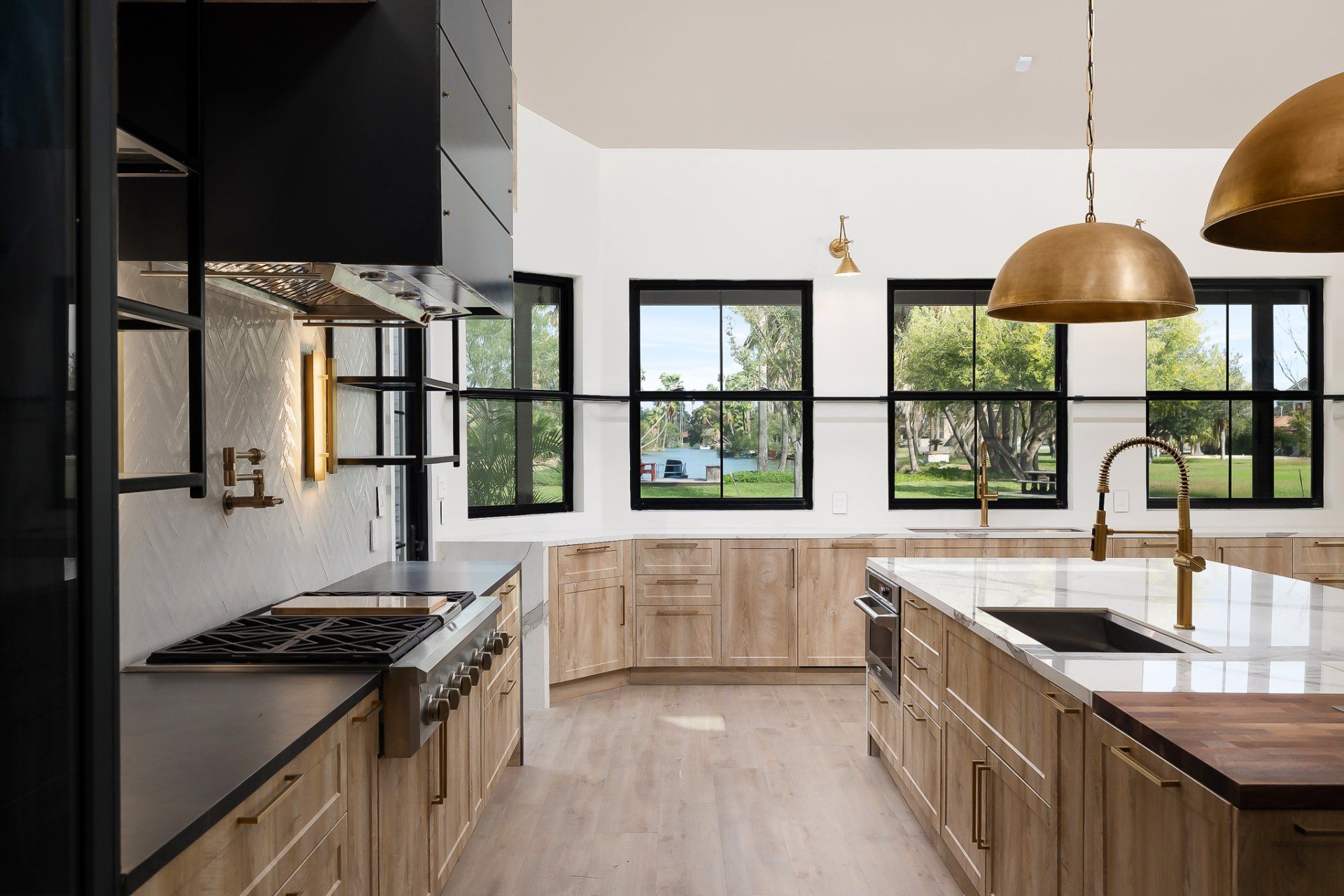 Modern bright kitchen with black and wood cabinets, white island, brass pendants, and large windows opening to greenery