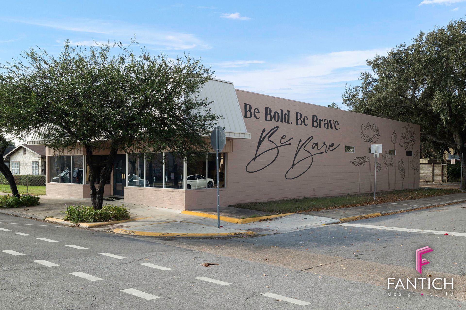Exterior of Be Bold Be Brave boutique with tan mural wall, trees, and parking lot in front