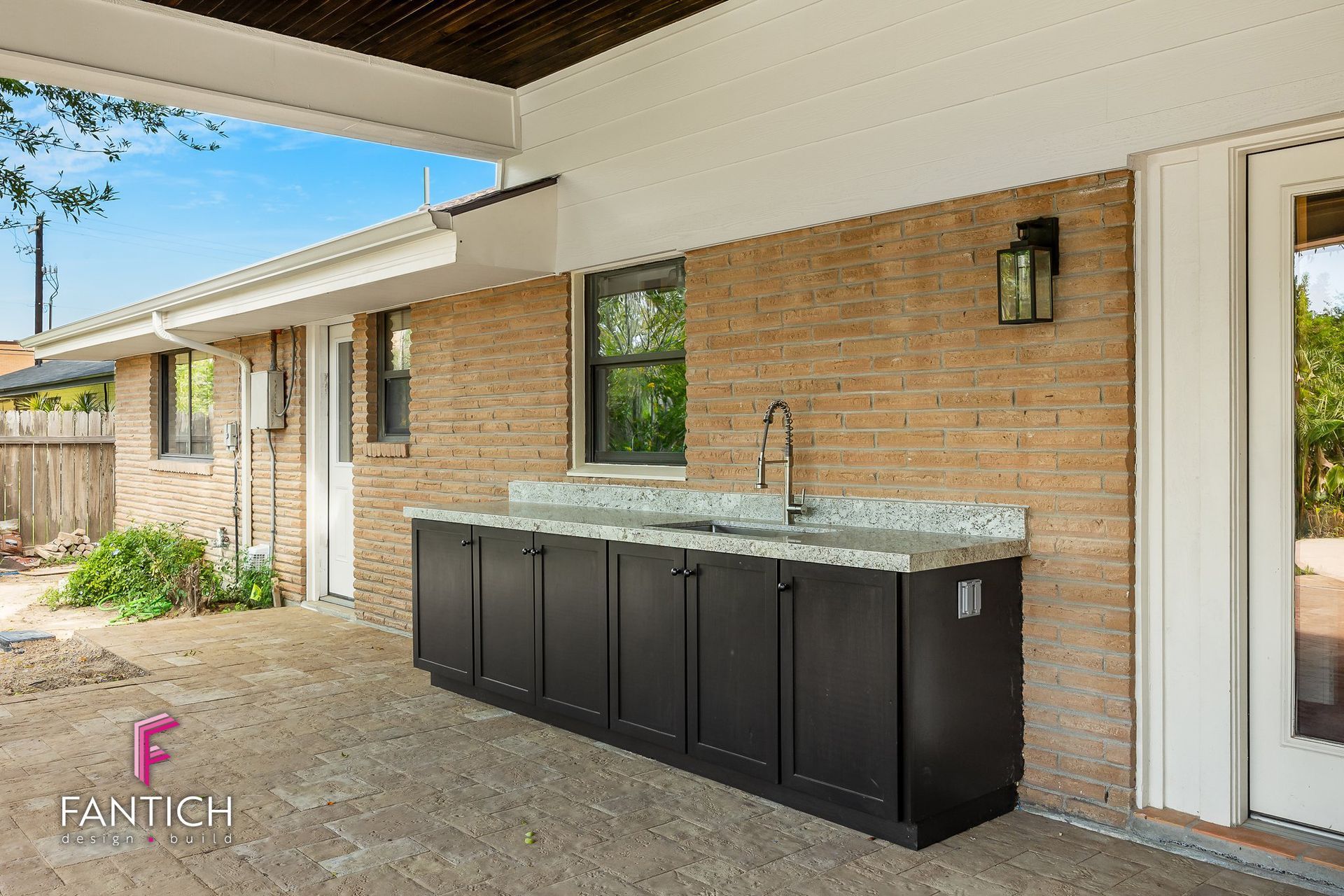 Covered patio with outdoor sink and black storage cabinets against a brick wall.