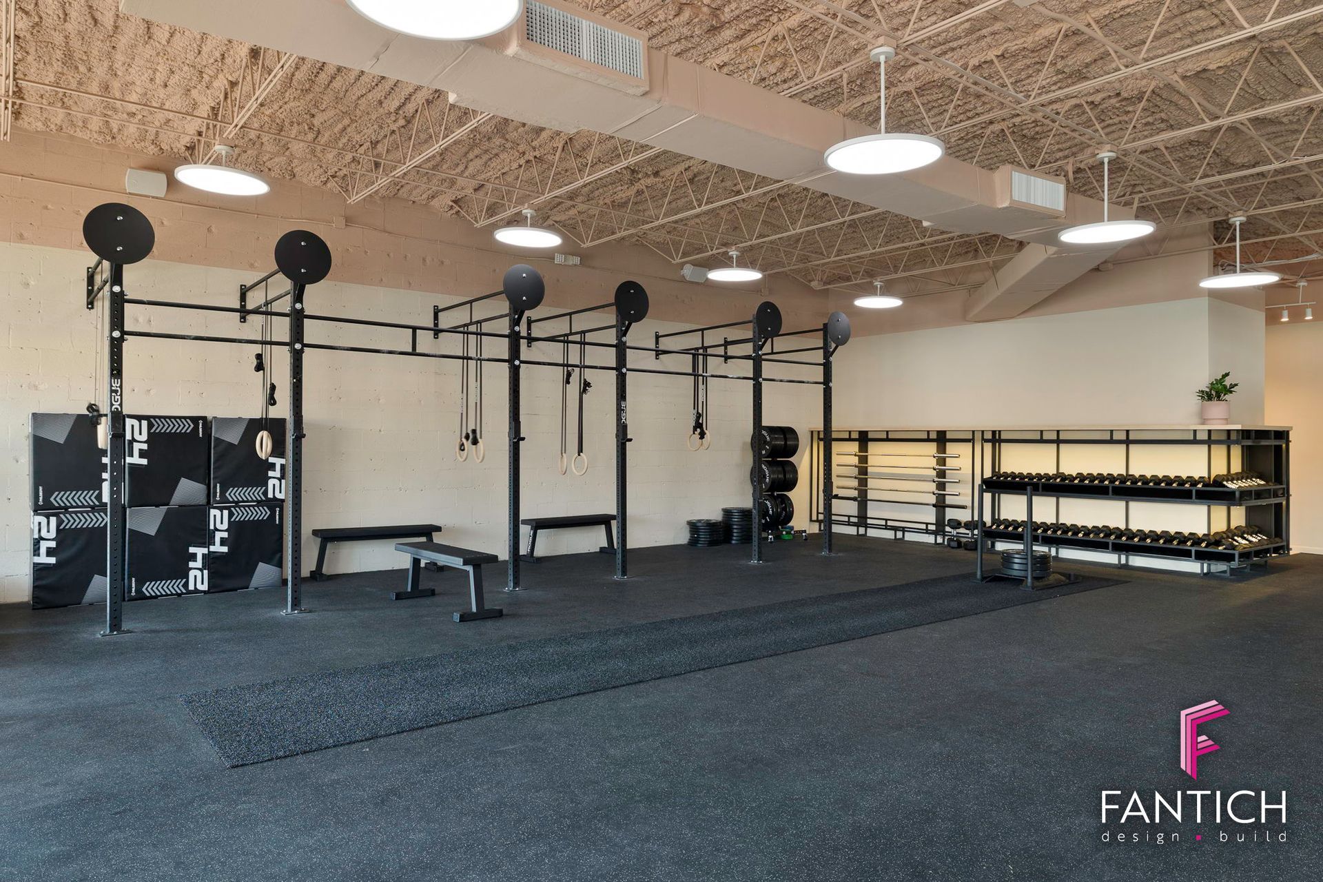 Empty gym with squat racks, dumbbells, and black flooring under exposed industrial ceilings.