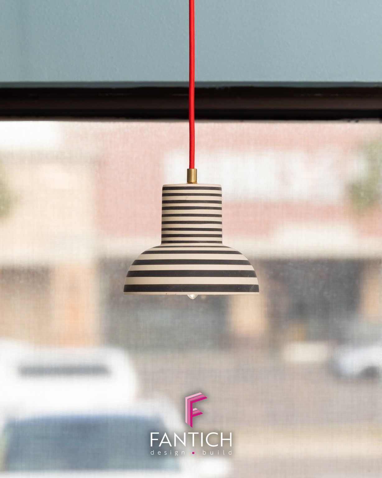 Striped pendant lamp with red cord hanging in front of a blurred window.