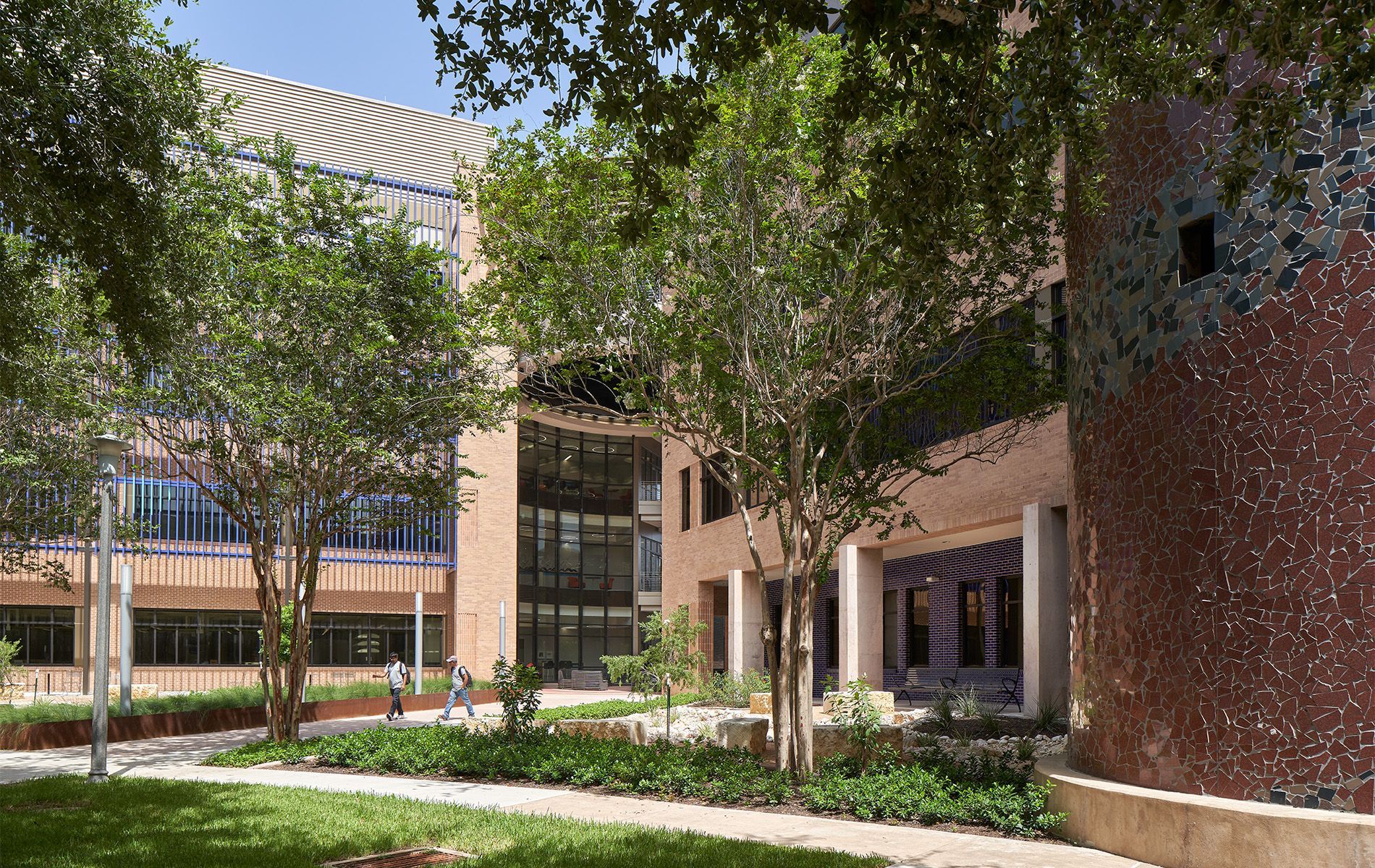 Sunlit courtyard with trees and brick campus buildings beside a glass walkway