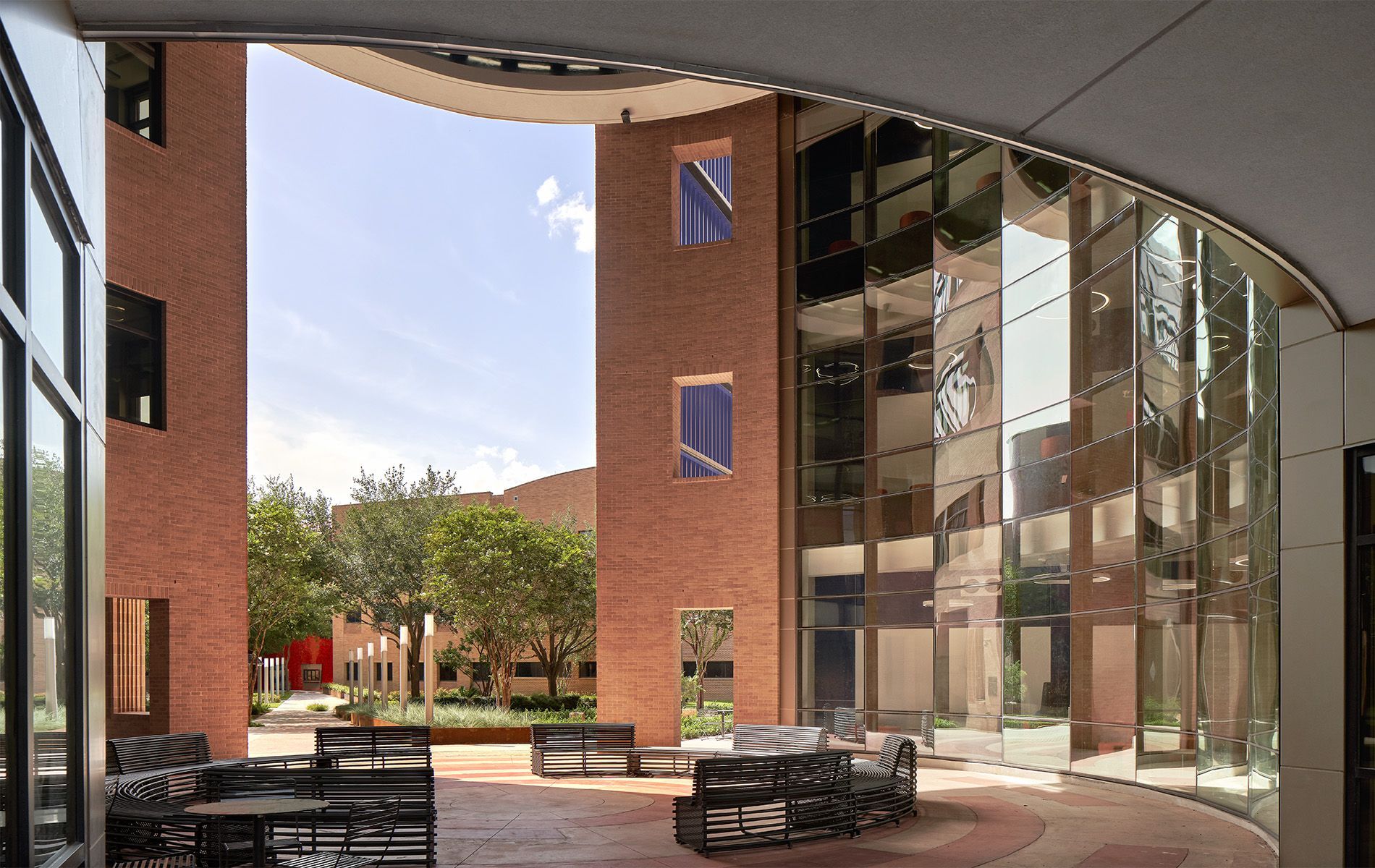 Modern courtyard with brick buildings, glass wall, and outdoor tables beneath a curved canopy