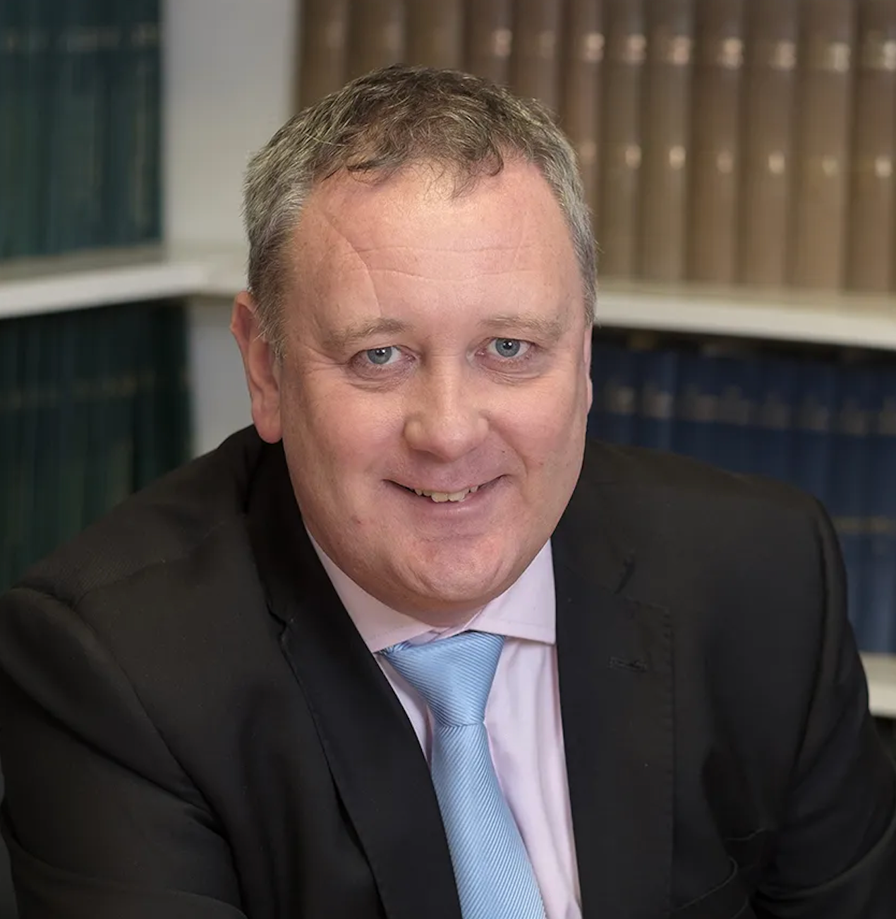 A man in a suit and tie is sitting in front of a bookshelf.