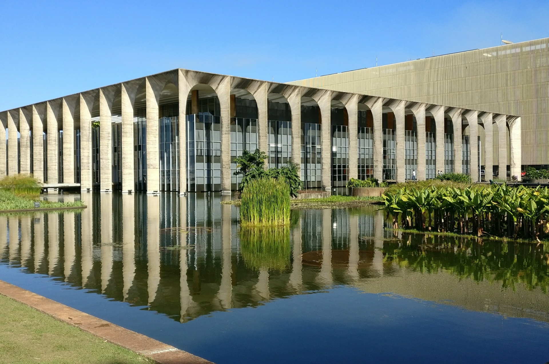 Edifício moderno de concreto com altas colunas refletido em um curso d'água tranquilo sob um céu claro.