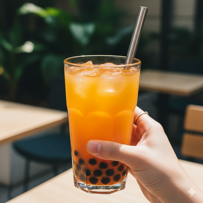 Una mano che regge un bicchiere di bubble tea arancione con ghiaccio e una cannuccia di metallo, all'aperto in un bar.