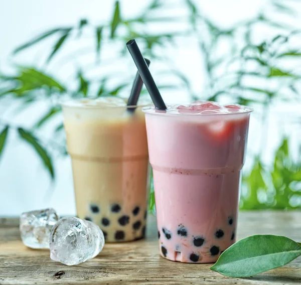 Due bevande al boba in bicchieri di plastica trasparente, uno beige e uno rosa, con cannucce nere. Cubetti di ghiaccio e vegetazione sullo sfondo.