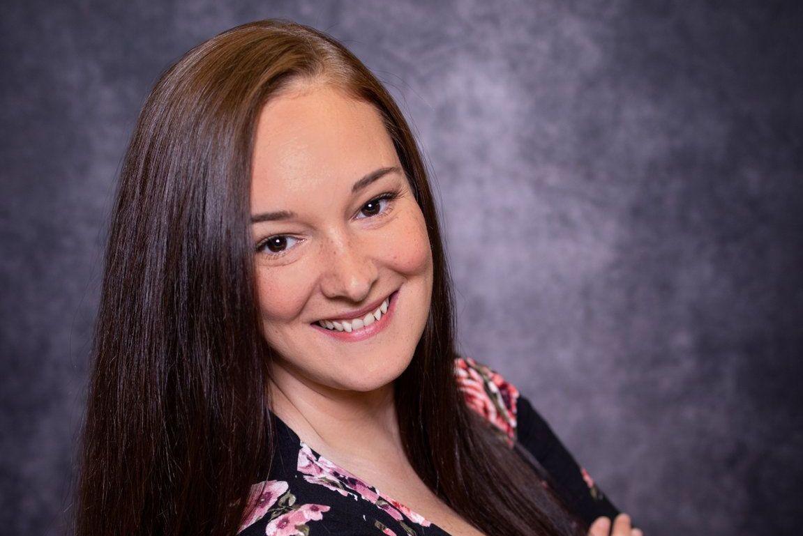 A woman with long hair is smiling for the camera in front of a gray background.