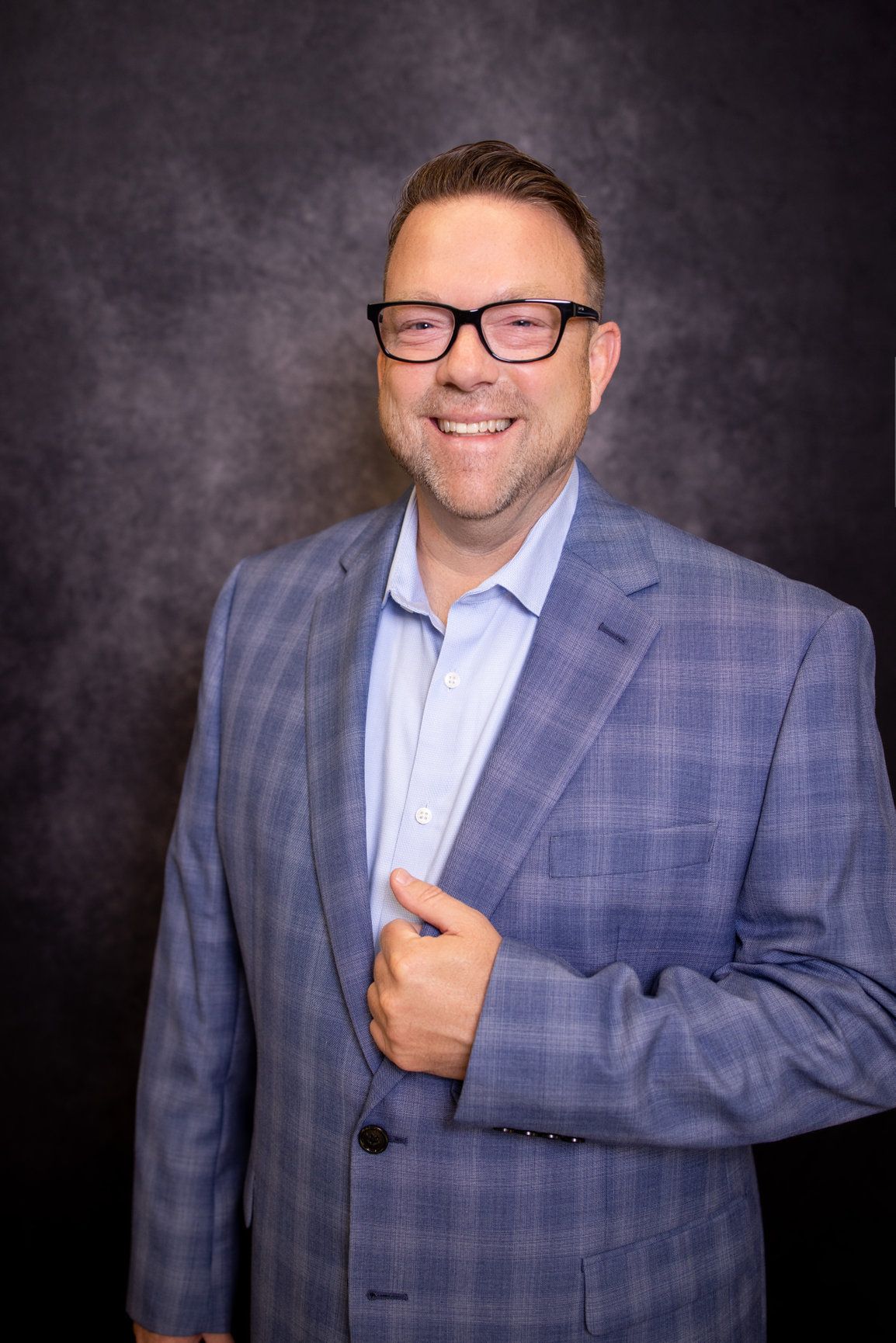 A man in a blue suit and glasses is smiling for the camera.