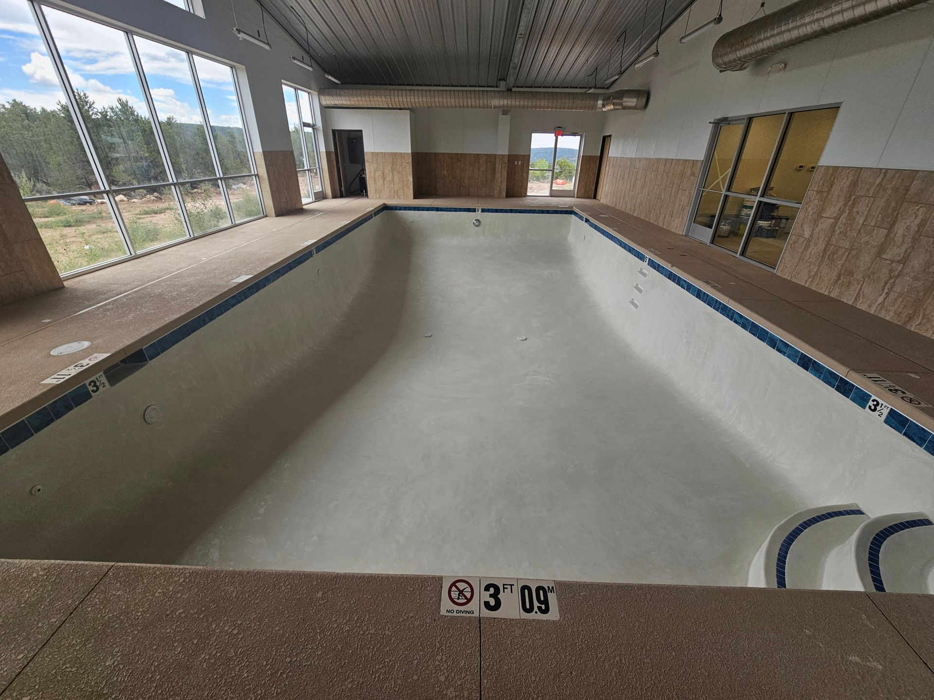 An unfinished concrete swimming pool in a backyard, featuring built-in stairs, a bench, and plumbing pipes.