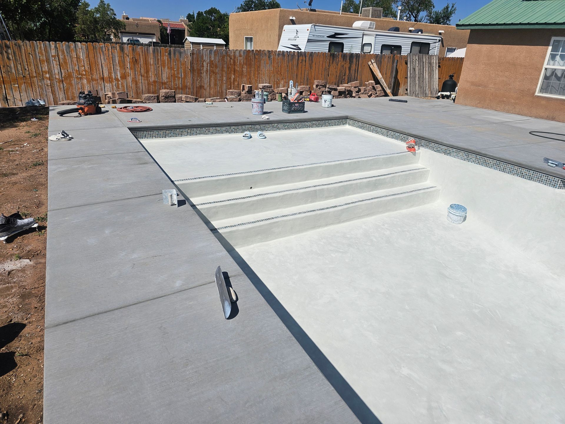 Freshly poured wet concrete surrounds an unfinished backyard swimming pool on a sunny day.