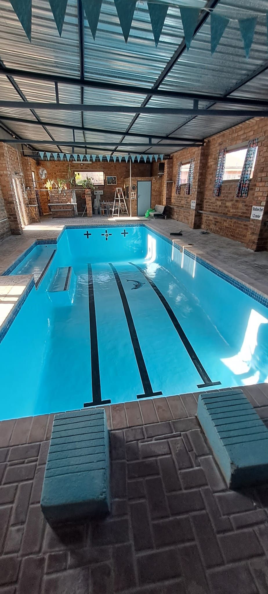 Indoor pool with blue water under a covered brick patio, featuring lane markings and lounge chairs nearby