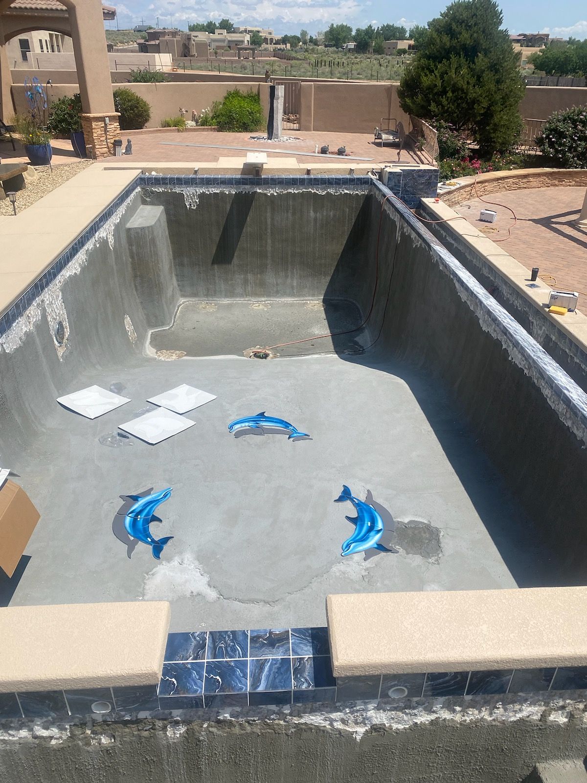 A backyard swimming pool with a circular brick-walled fountain feature in the foreground and a gray house in the back.