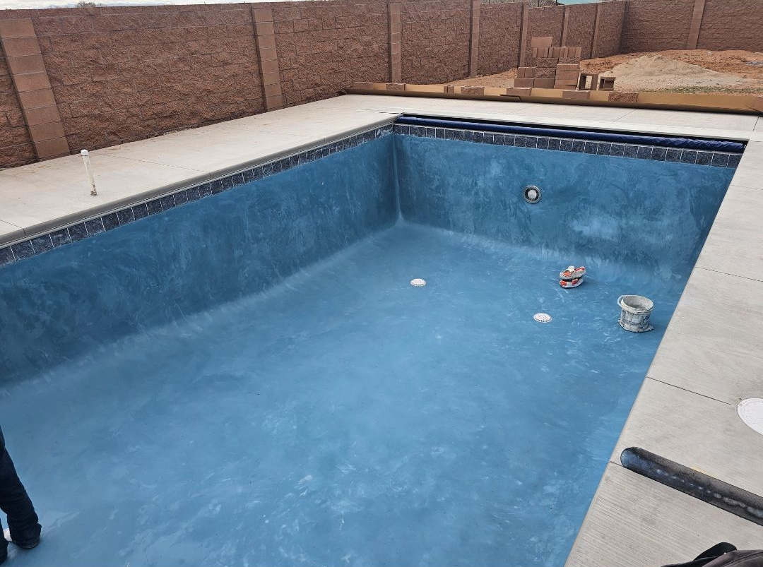 An empty, bright blue swimming pool sits surrounded by stone pavers in a sunny backyard setting.