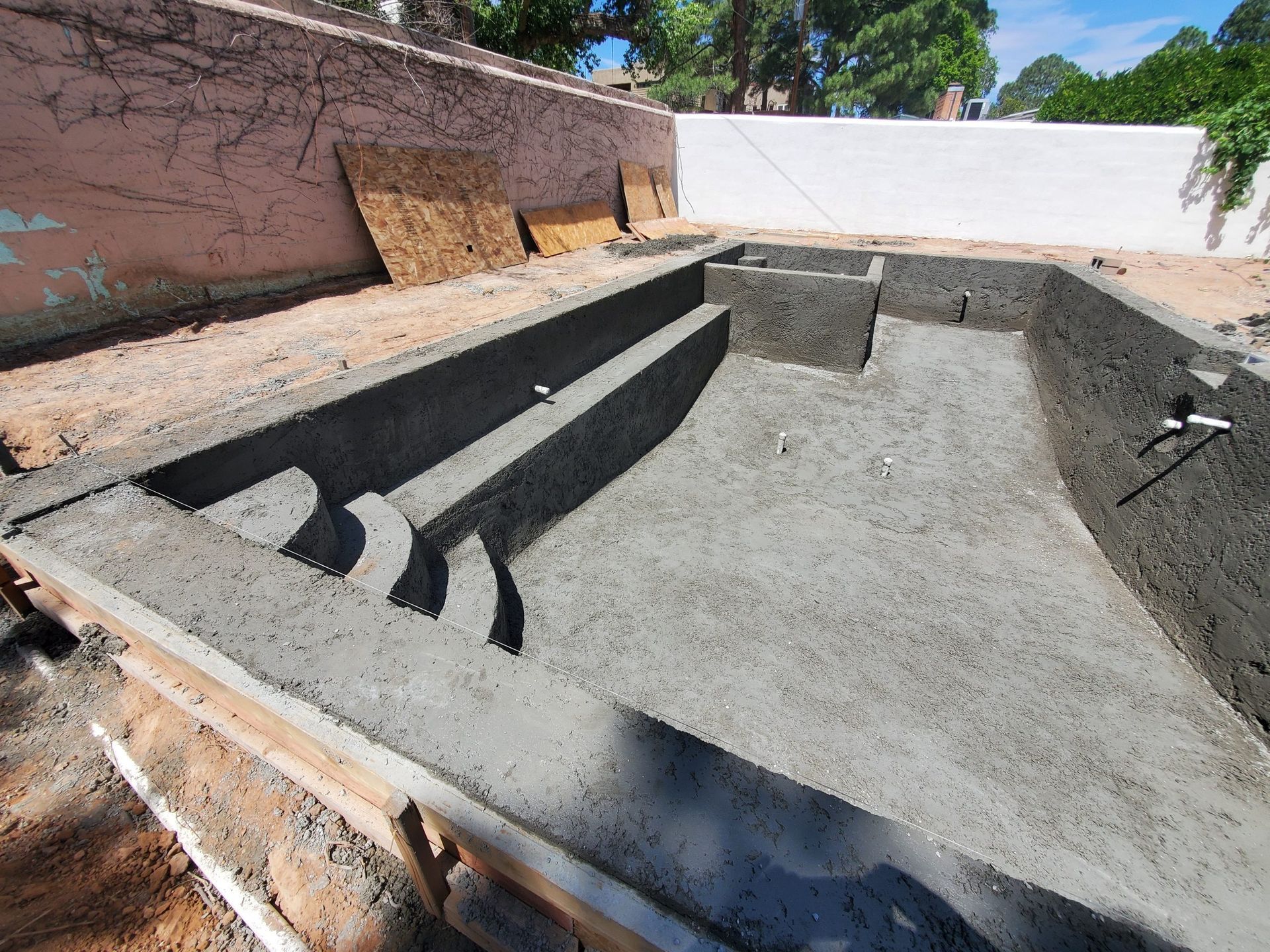 Empty concrete swimming pool under construction with stairs and built-in bench, surrounded by dirt and walls