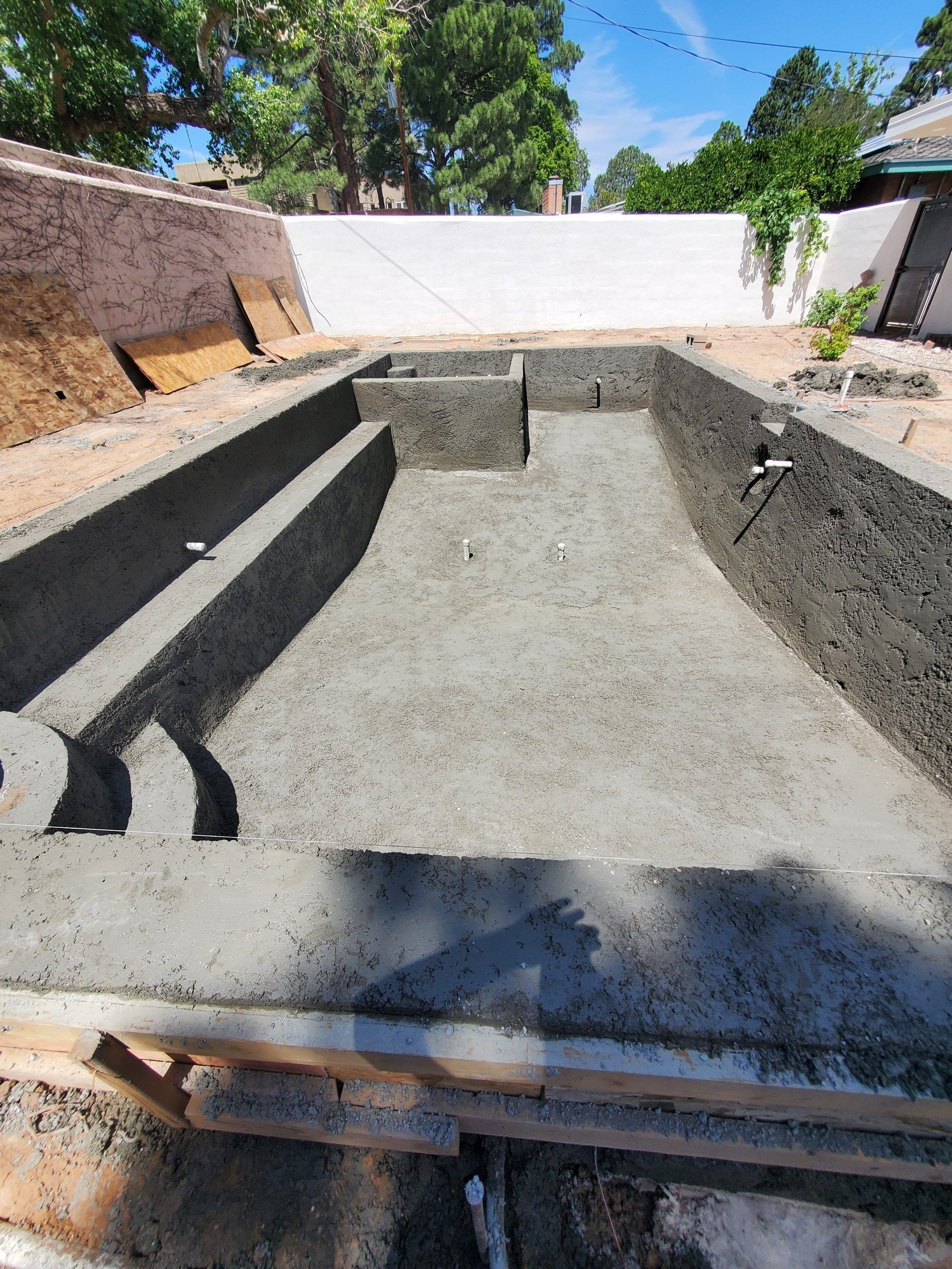 Empty concrete swimming pool under construction in a backyard, with blue sky and trees above.