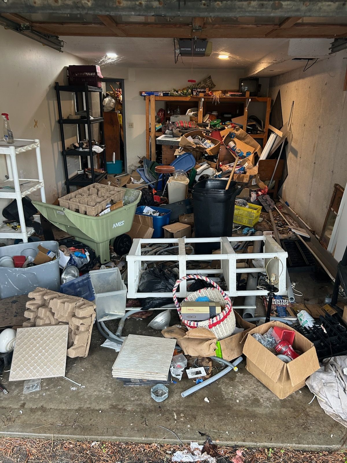 Cluttered garage filled with boxes, bins, and scattered household items, with shelving and a car partially visible.