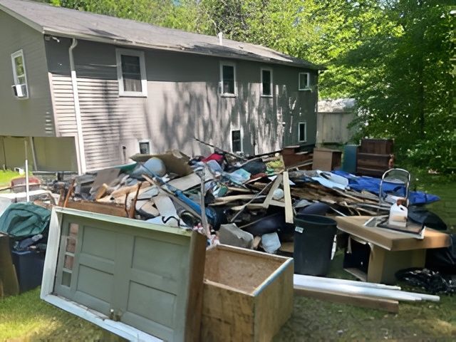 Pile of junk in a backyard. Gray house in the background. Includes wood, trash bags, and furniture.
