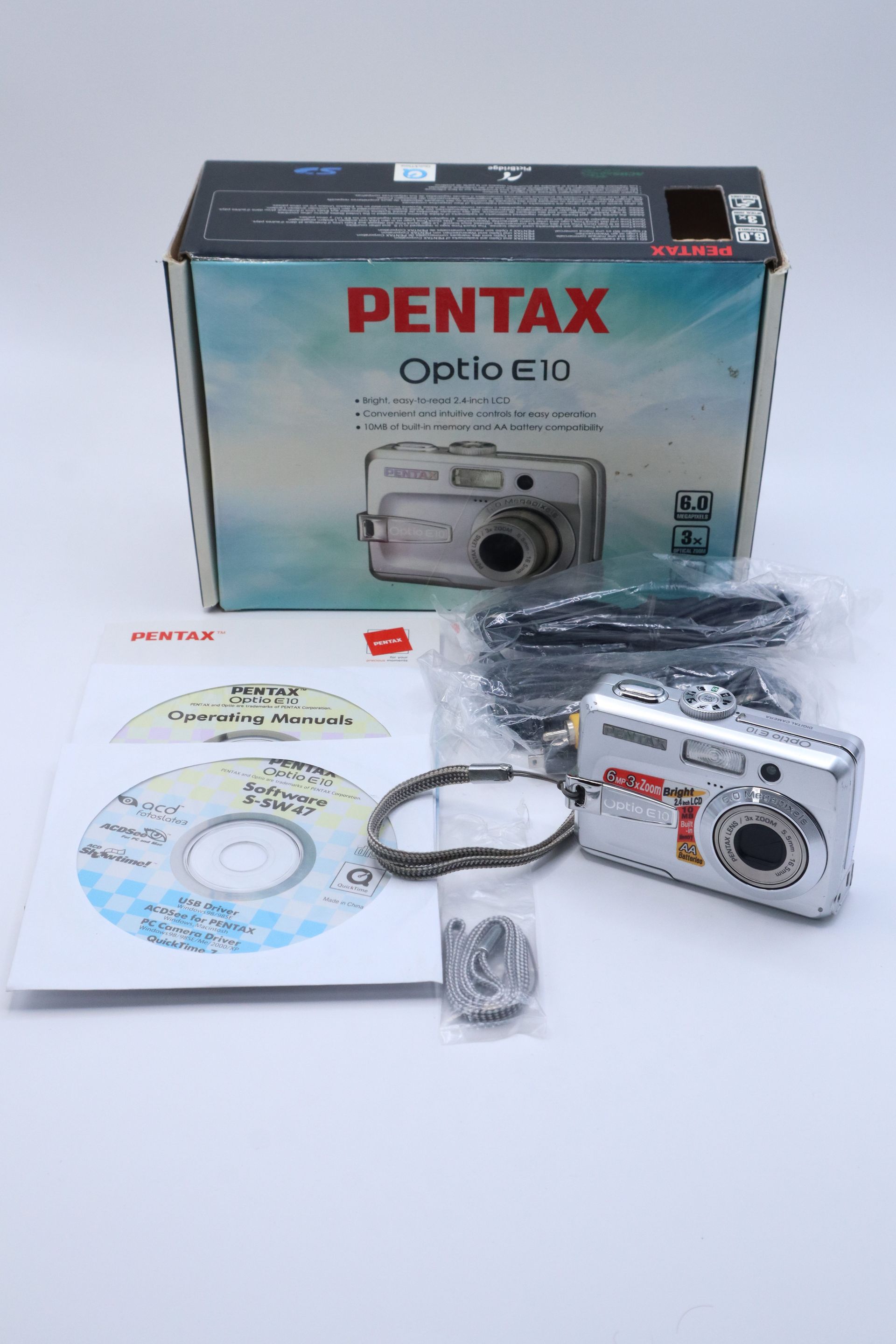 Pentax Optio E10 camera box with silver digital camera, CD, cable, and accessories laid out on a white surface