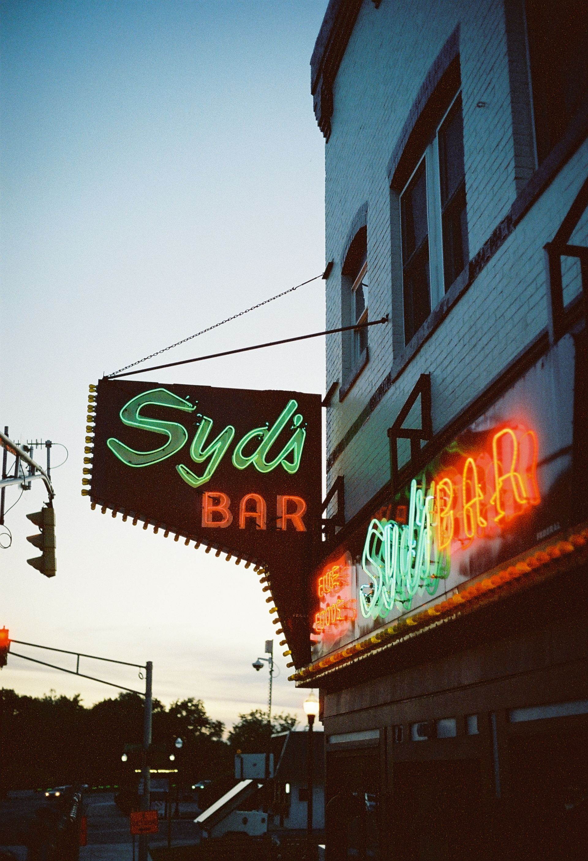 Neon signs for Syd’s Bar and a red-orange storefront glow on a dusk street corner