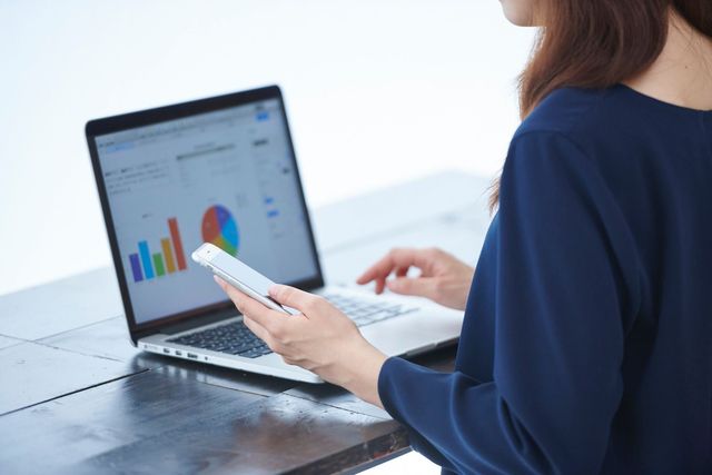 Woman using laptop and smartphone, analyzing charts and graphs on a light-colored desktop.