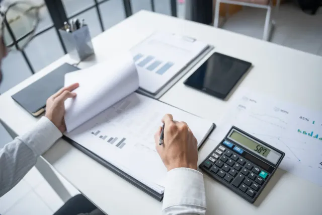 Hands reviewing financial documents with a calculator, tablet, and pen on a white desk.
