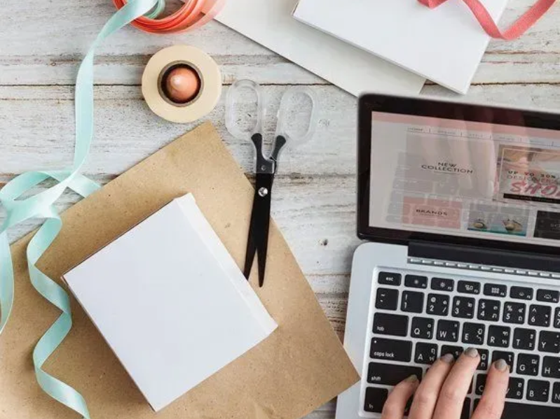 Desk with notebook, tape, ribbons, scissors, and laptop beside hands typing on a wooden workspace