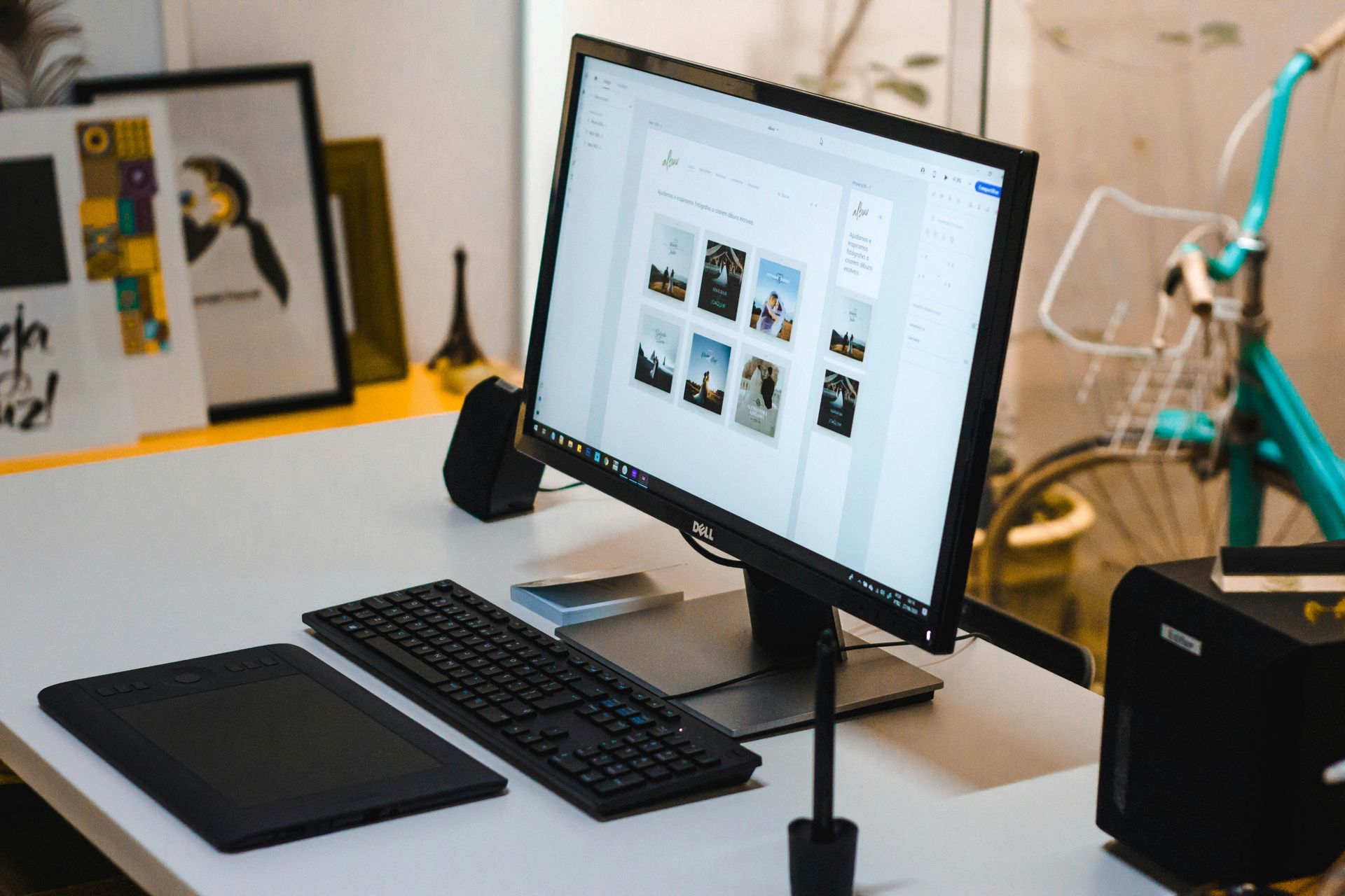 Desk with a monitor showing a photo gallery, keyboard, drawing tablet, and speakers in a bright workspace.