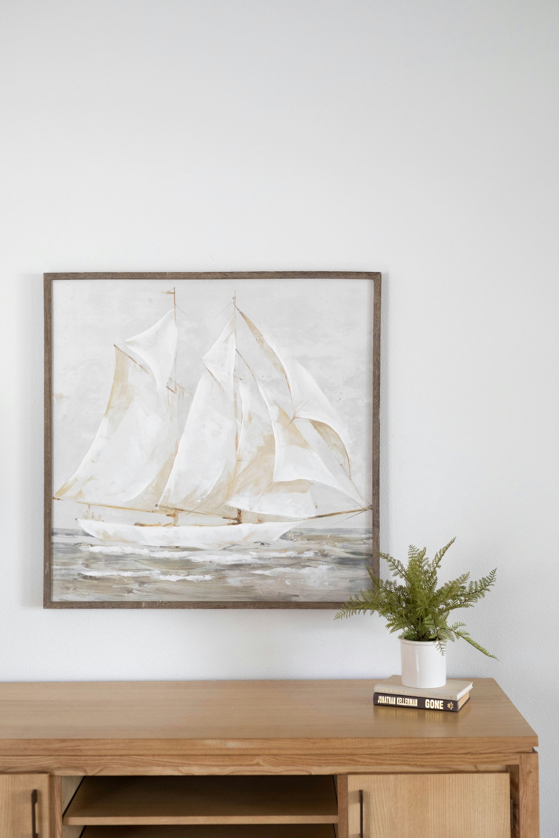 Framed white sailboat painting above a wooden console with a small potted plant and books