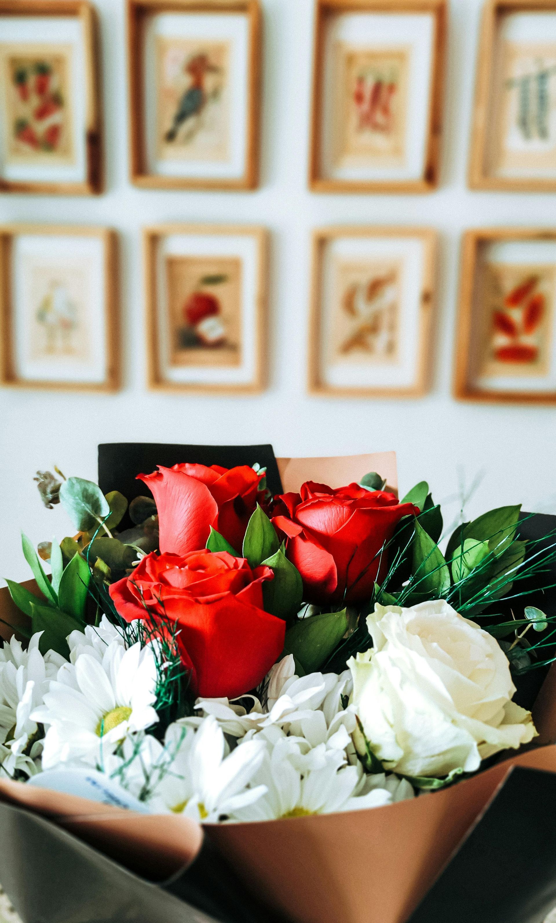 Bouquet of red, white, and green flowers in paper wrap, with framed artwork on the wall behind