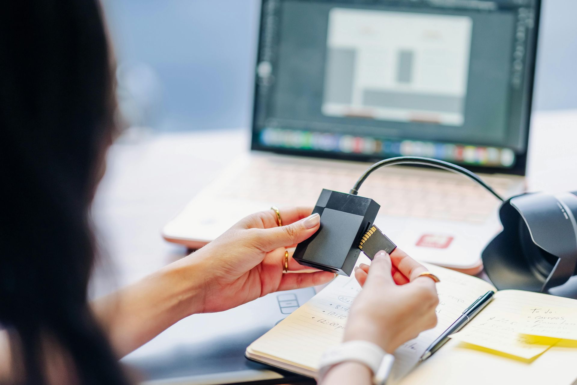 Hands holding a smartphone over a notebook beside a laptop on a desk