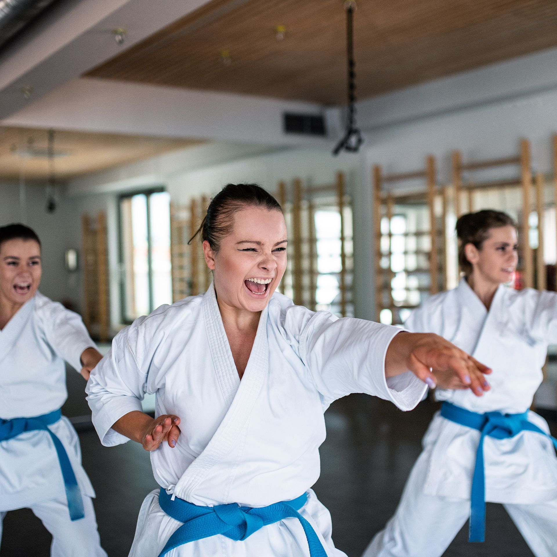 Three women in white karate uniforms with blue belts are practicing karate in a gym.