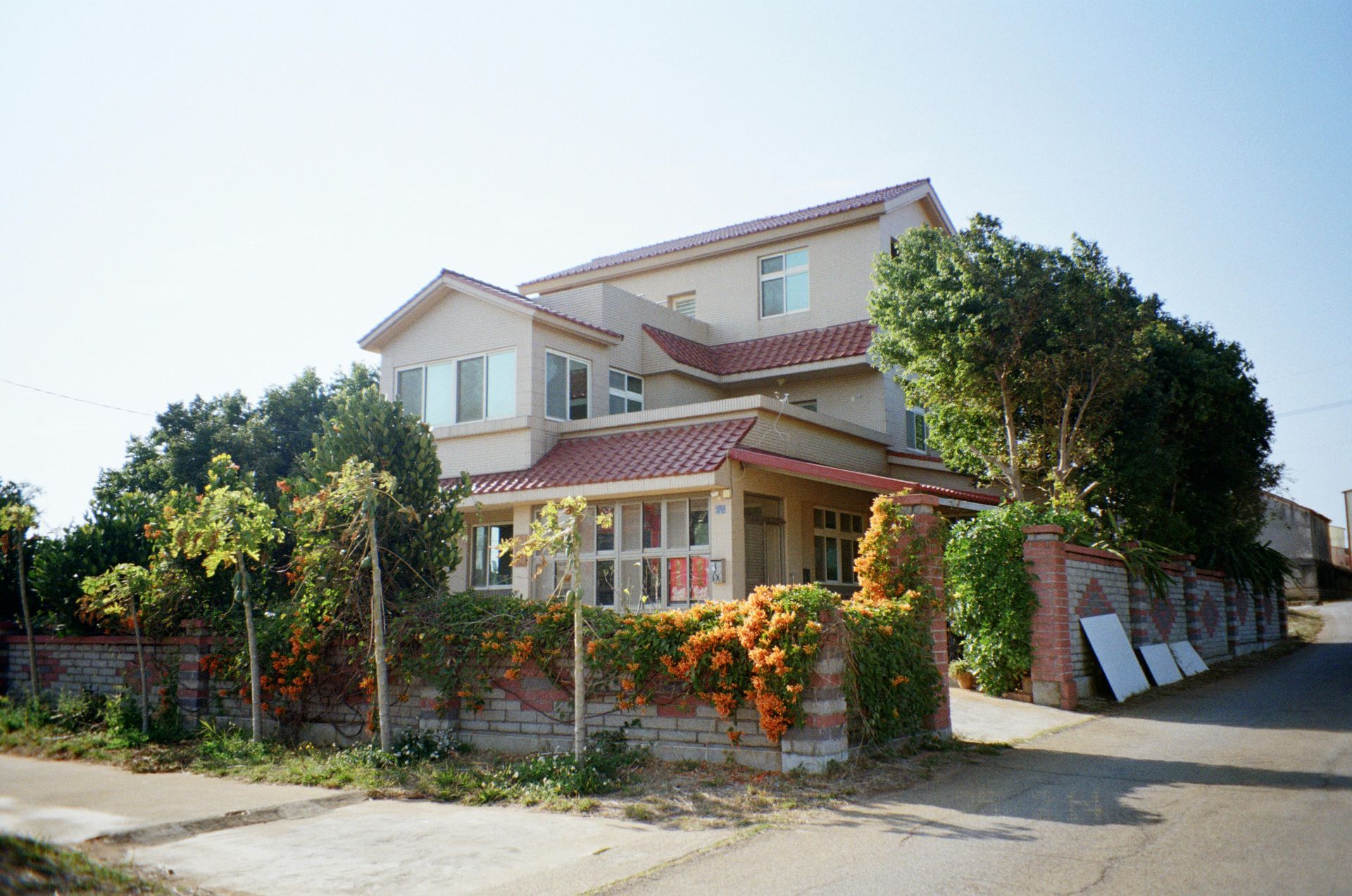 A large house with a red tile roof is surrounded by trees and bushes.