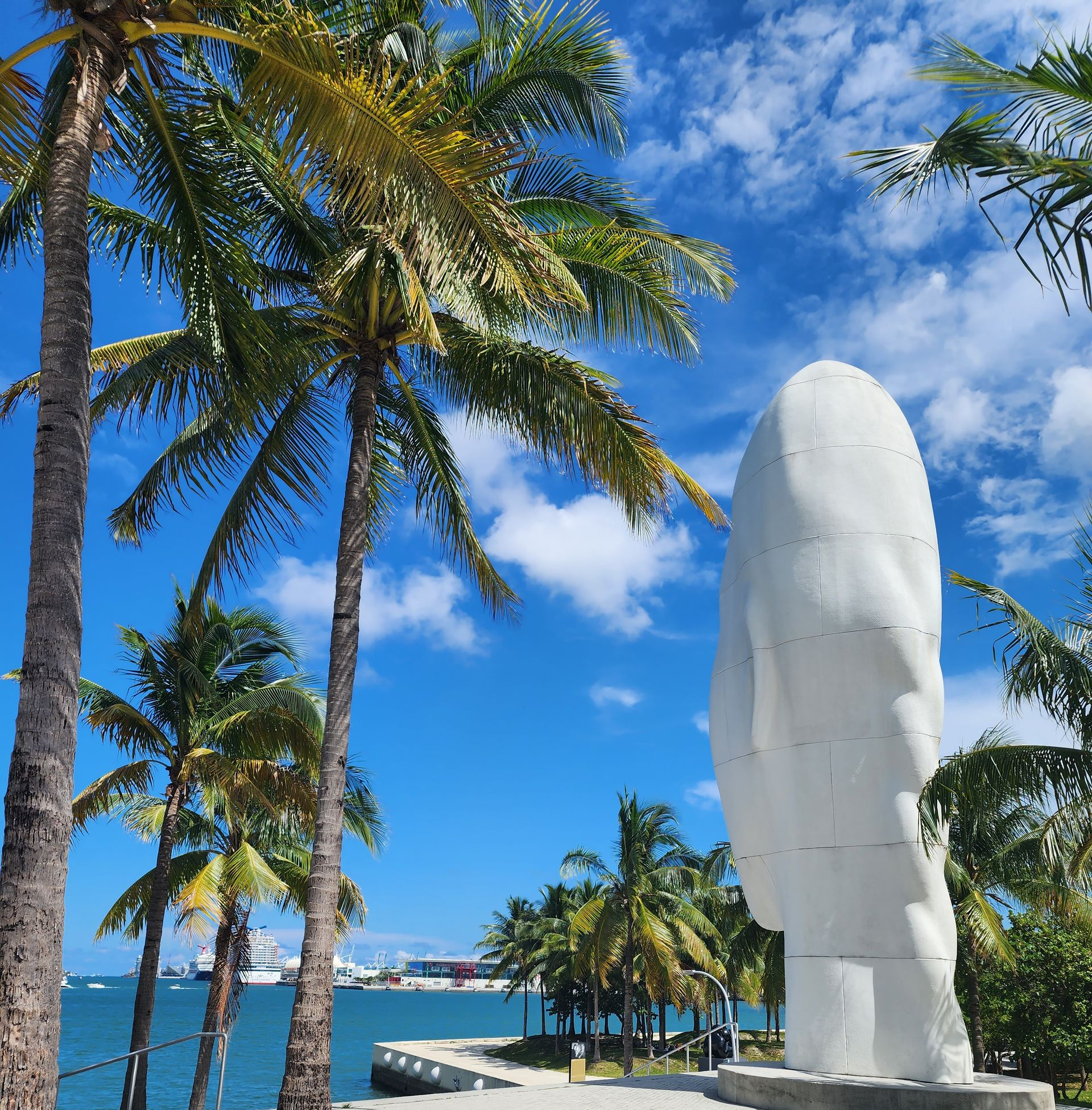 Tropical waterfront with palm trees, blue sky, and a tall white sculpture by the shore