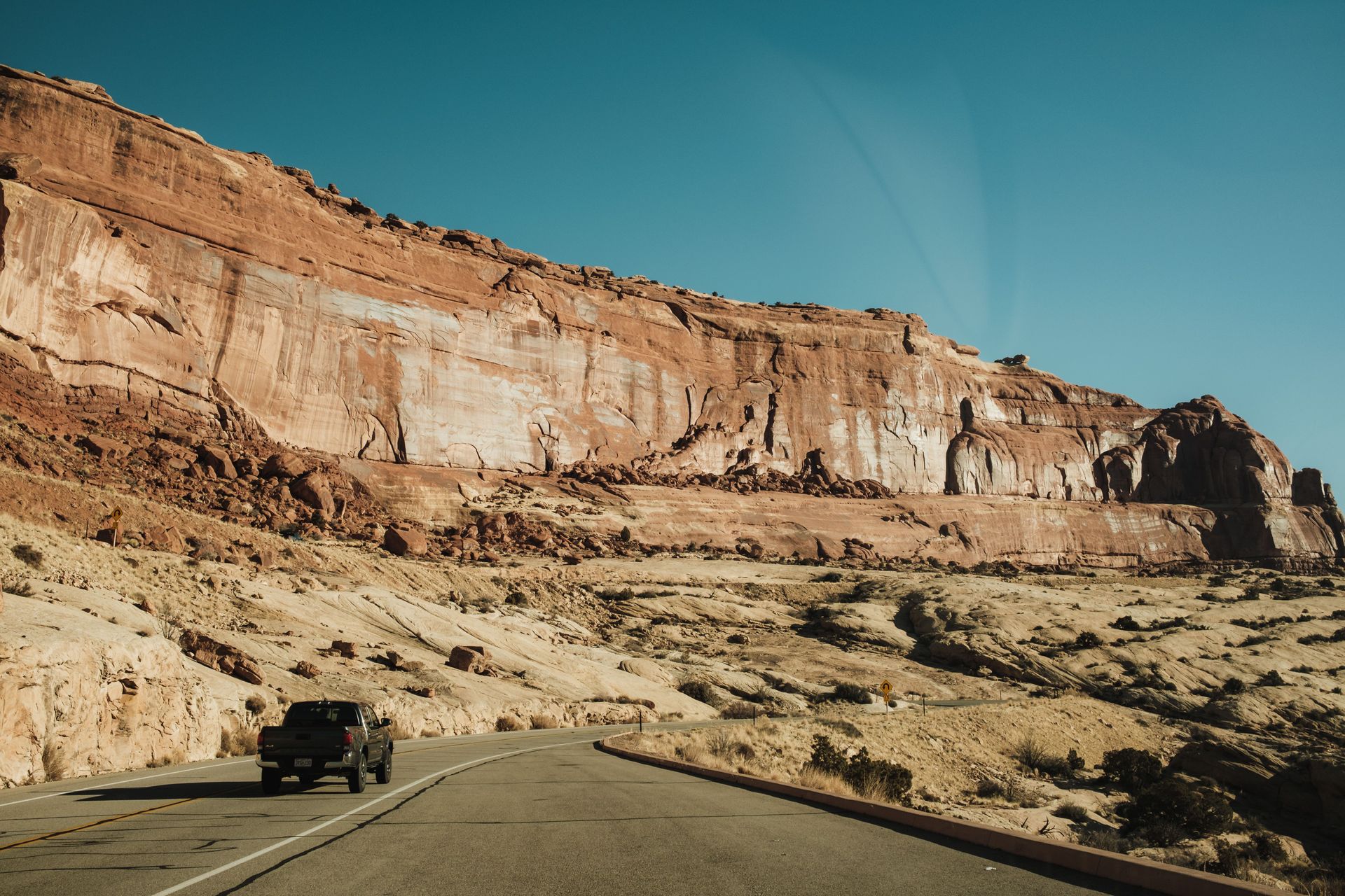 Car on a desert road beside a towering red rock cliff under a clear blue sky