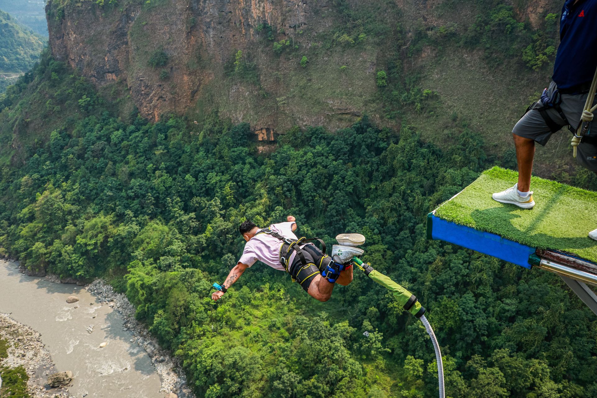 Person bungee jumping from a platform over a green canyon with a river below