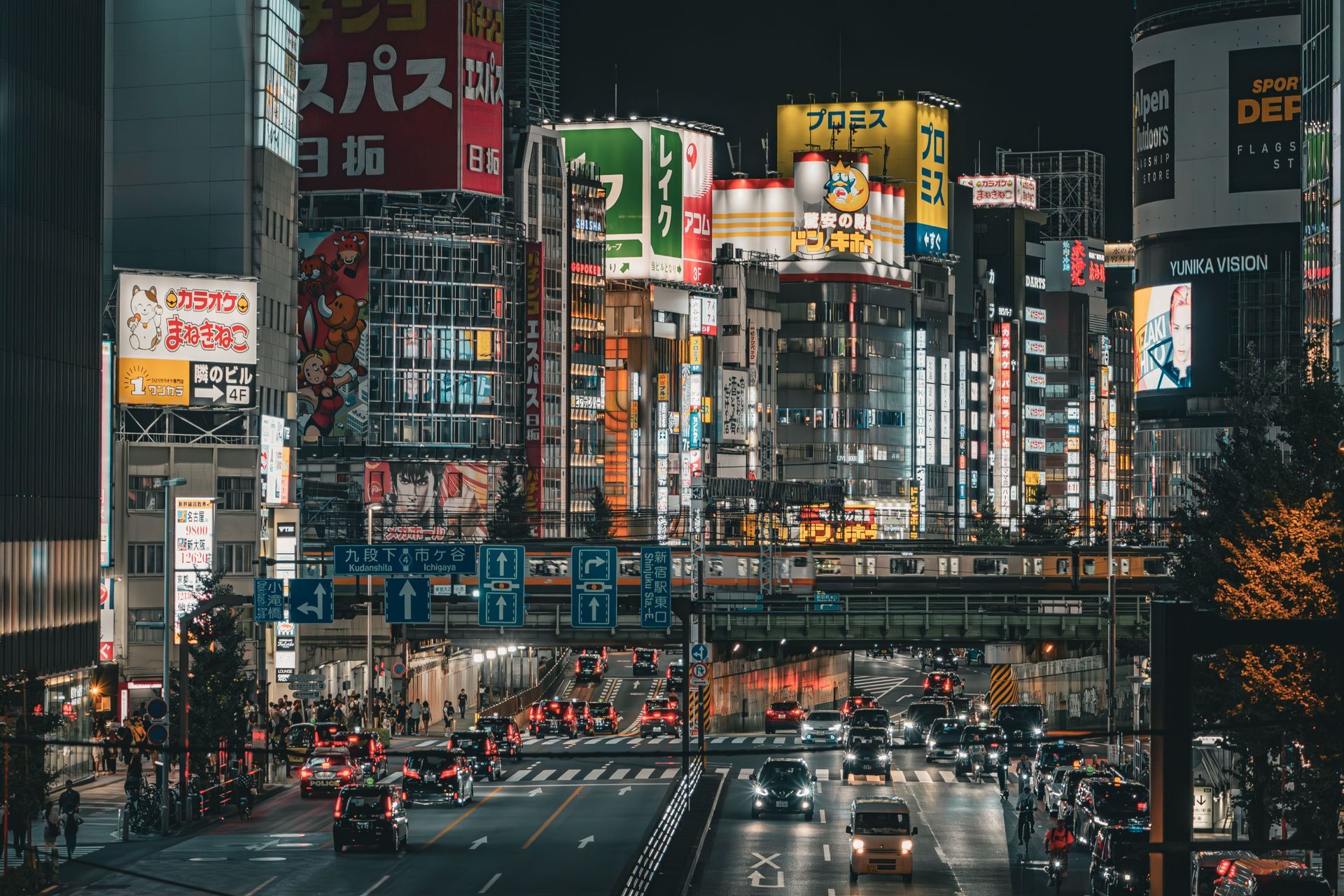 Busy Tokyo street at night with neon billboards, elevated train tracks, and traffic below