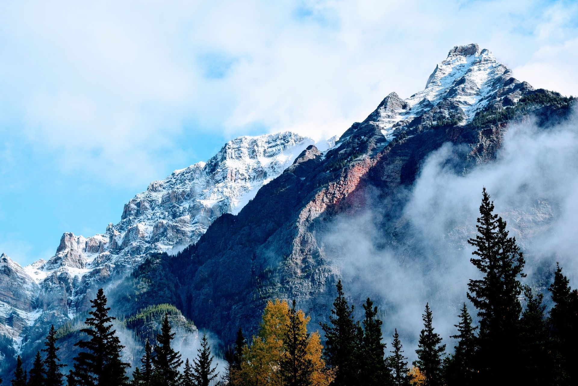 Snowy mountain peaks above dark pine trees under clouds and blue sky