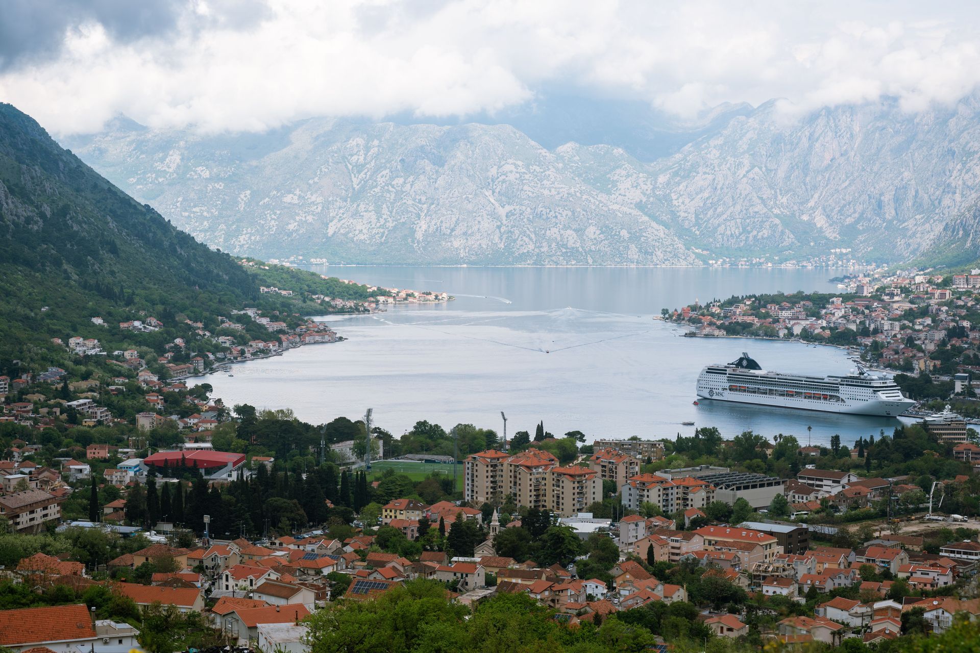Coastal town with red-roofed buildings, a calm bay, and mountains in the misty background.