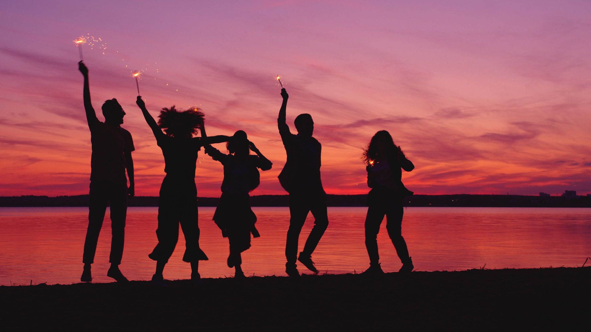 Five silhouetted people dancing at sunset by a calm lake with a pink-purple sky