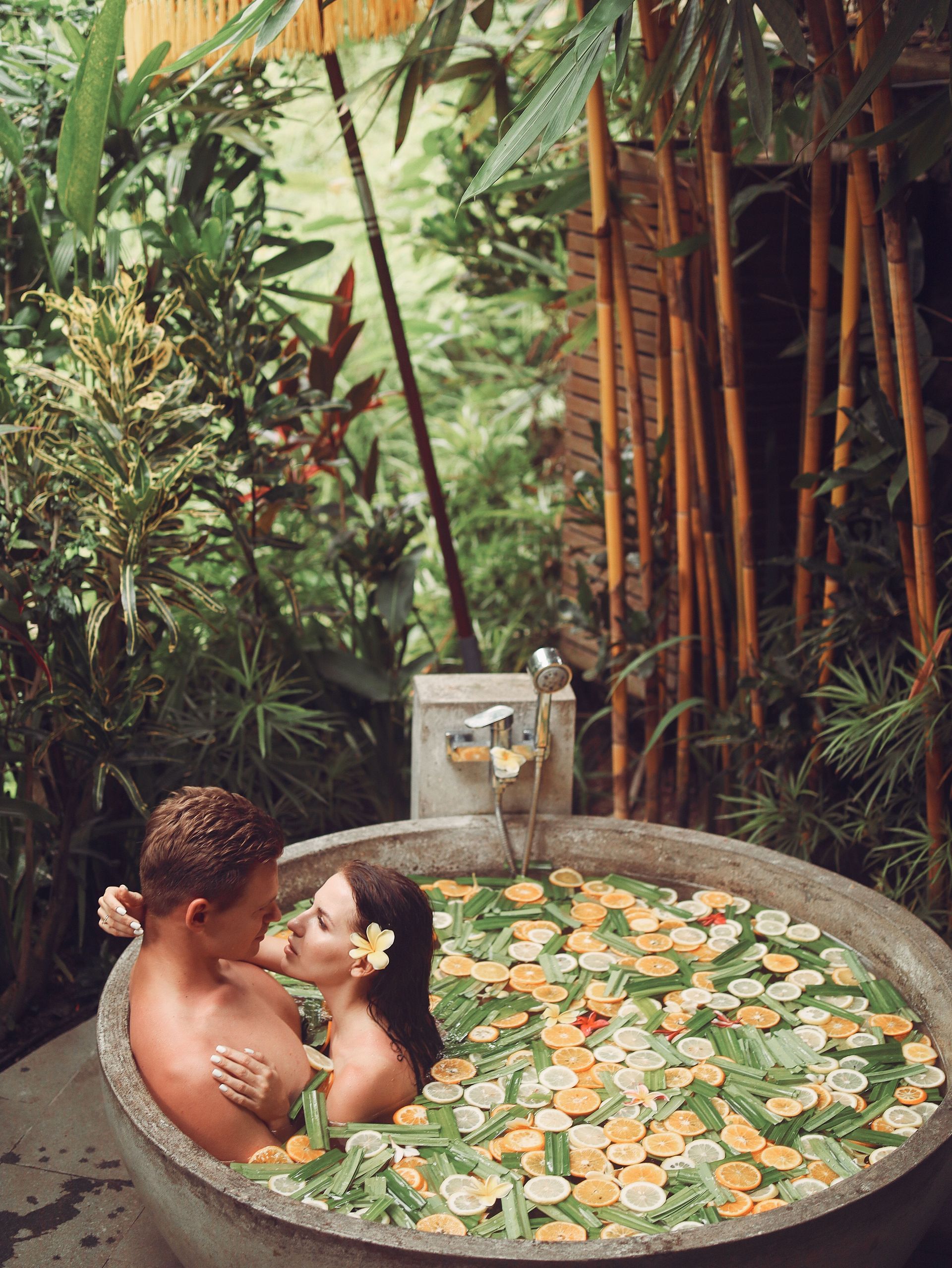 Couple relaxing in a round stone hot tub surrounded by tropical plants and floating flower petals.