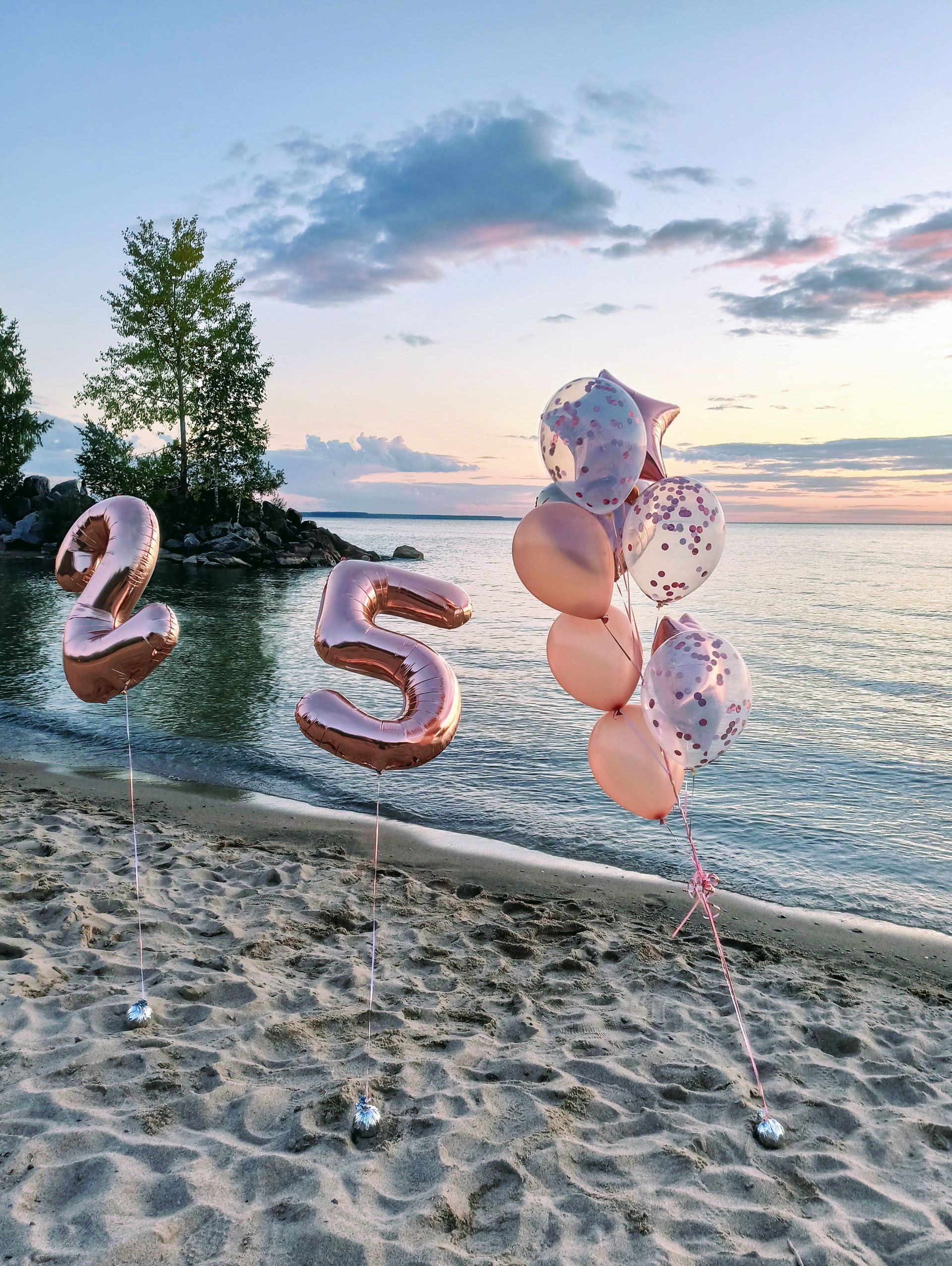 Rose gold number balloons on a sandy beach at sunset with water and trees in the background