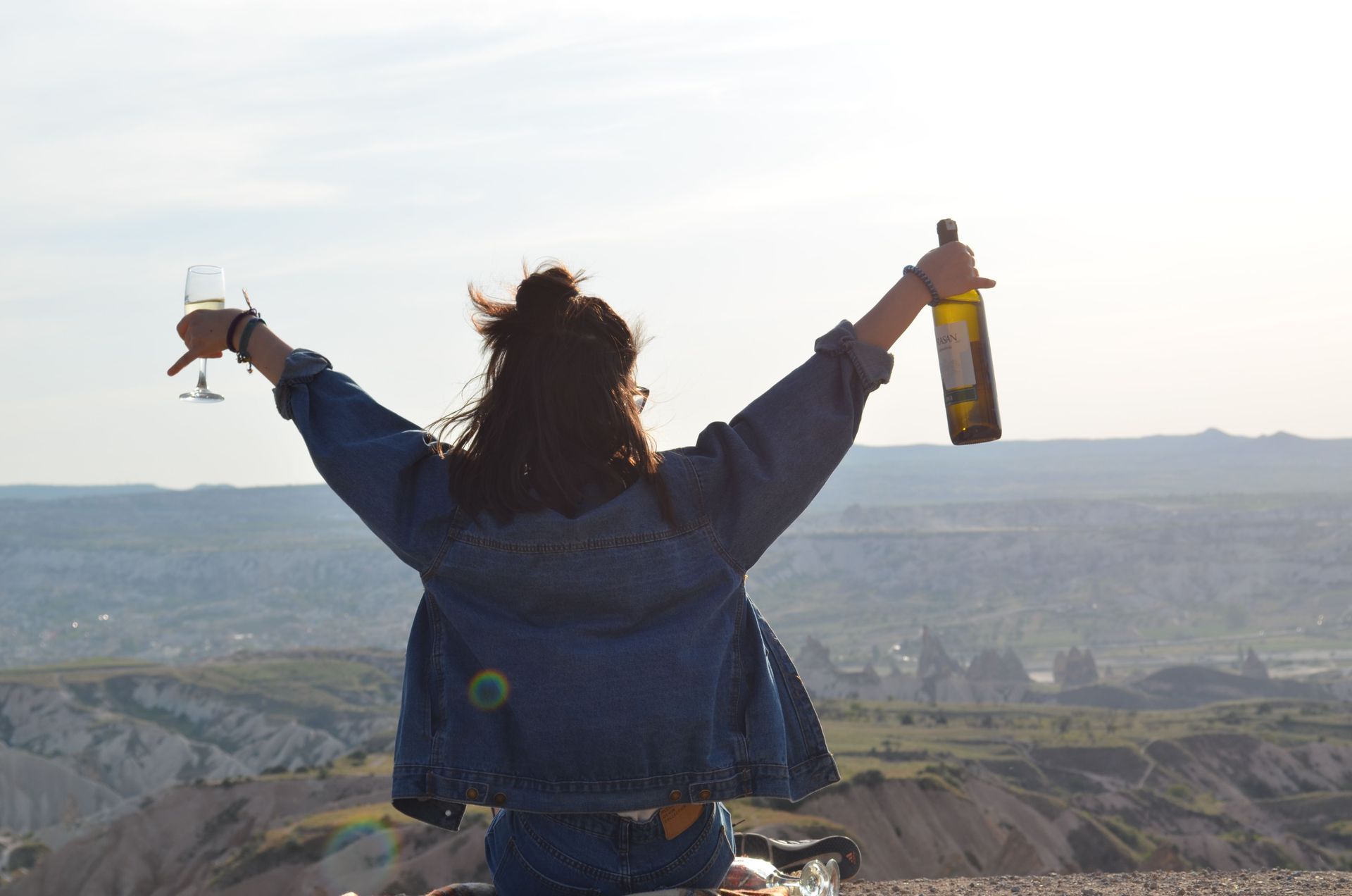Person with arms raised holding a wine glass and bottle, overlooking a scenic valley at sunset