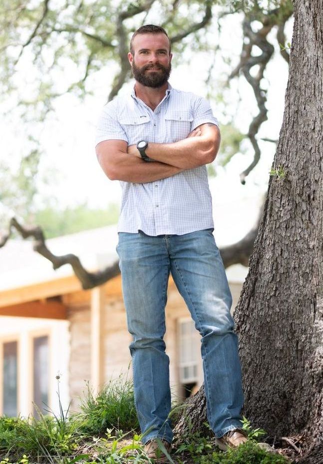 A man with a beard is standing next to a tree with his arms crossed.