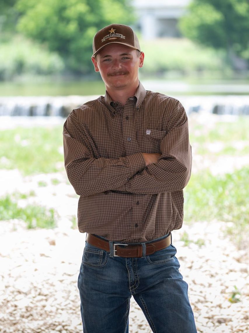 A man wearing a hat and a brown shirt is standing with his arms crossed.